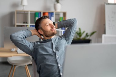 Rahat, olgun bir şirket çalışanı sandalyeye yaslanmış, elleri başının arkasında, iş yerinde işini bitirdikten sonra dinleniyor.