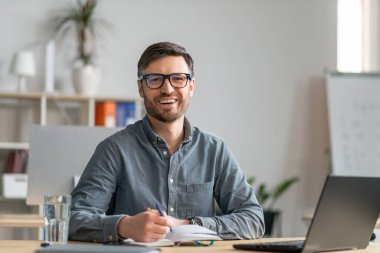 Mutlu yetişkin girişimci ofiste online eğitim alıyor, dizüstü bilgisayar kullanıyor, notlar alıyor ve kameraya gülümsüyor, boşluğu kopyalıyor.