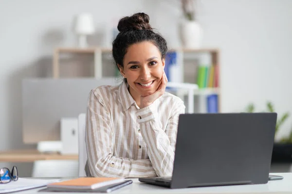 Портрет счастливой деловой женщины, сидящей на рабочем месте перед ноутбуком и улыбающейся перед камерой в современном офисе — стоковое фото