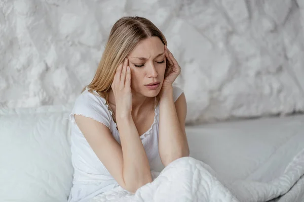 Verdrietig jong europees blond vrouw wakker, lijdt aan hoofdpijn in wit bed in slaapkamer — Stockfoto