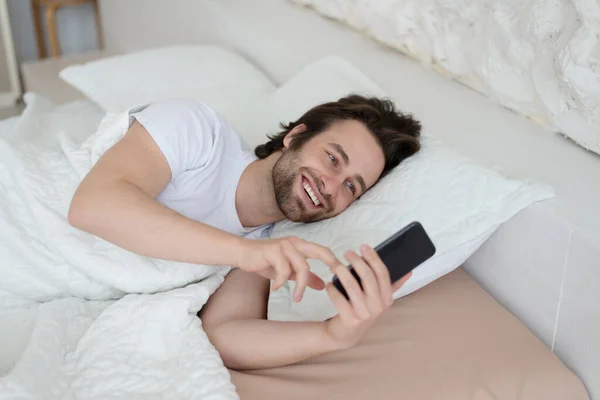 Sonriente joven europeo macho con rastrojo se despierta en la cama, charlando en el teléfono inteligente, leyendo noticias en el dormitorio — Foto de Stock