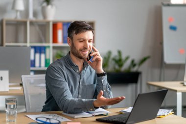 Mutlu bir iş adamı cep telefonuyla konuşuyor, iş sorularını tartışıyor, laptopa bakıyor, iş yerinde oturuyor.