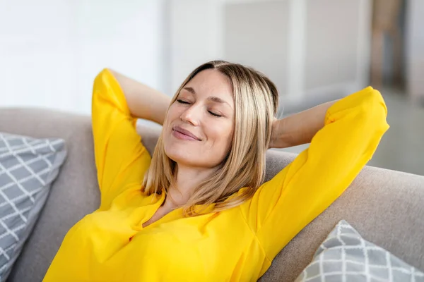 Feliz joven rubia europea en ropa amarilla con los ojos cerrados descansando en el sofá en el interior de la sala de estar — Foto de Stock