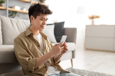 Gülümseyen Asyalı adam evde cep telefonu kullanıyor ve yerde oturuyor.