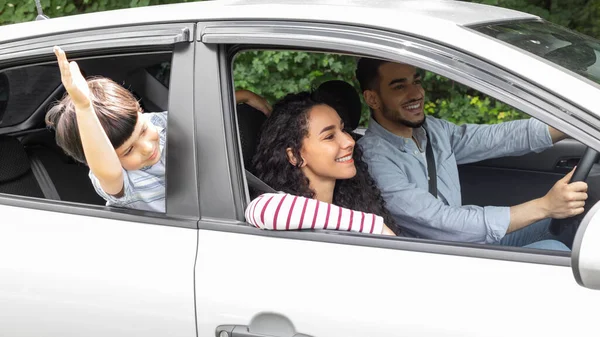 Feliz millennial Oriente Medio macho y hembra conduciendo en coche, niño pequeño saludando de la mano en la ventana abierta, al aire libre —  Fotos de Stock