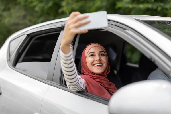 Gelukkig jong Arabisch dame in hijab neemt selfie op telefoon in open raam in auto, echtgenoot rijden, buiten — Stockfoto