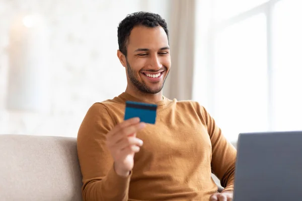 Hombre sonriente usando PC y tarjeta de crédito sentado en el sofá — Foto de Stock