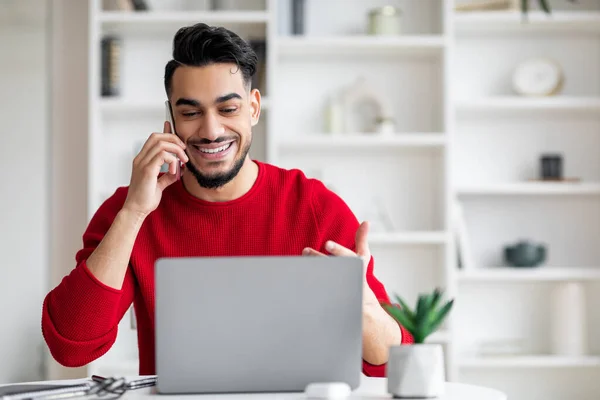 Usmívající se atraktivní tisíciletý arabský samec s plnovousem v červeném oblečení mluví telefonicky a dívá se na notebook — Stock fotografie