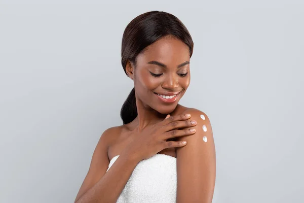 Cheerful pretty young black female in towel after shower applies cream to her shoulder, isolated on gray background — Stock Photo, Image