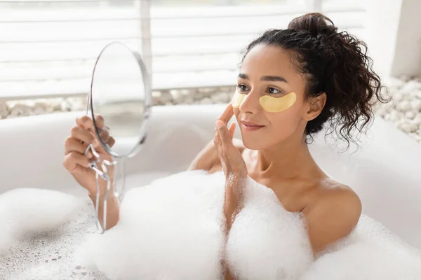 Ładna młoda kobieta nakładająca złote plamy pod oczami, patrząc w lustro leżąc w gorącej kąpieli bąbelkowej w domu — Zdjęcie stockowe