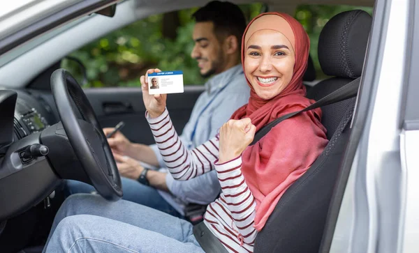 Blij moslim vrouw in hijab tonen rijbewijs en vuist in open deur in auto na examen met mannelijke leraar — Stockfoto