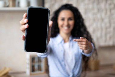 Mutlu kadın boş ekranlı akıllı telefonu gösteriyor ve onu işaret ediyor, mutfağın içinde duruyor, seçici bir odak noktası var.