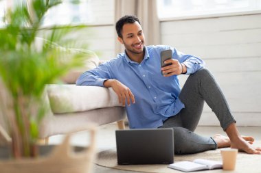 Evde akıllı telefon ve bilgisayar kullanan gülümseyen bir adamın portresi