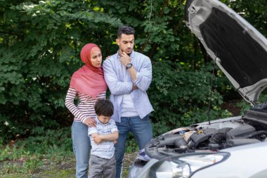 Üzgün, düşünceli Müslüman adam, tesettürlü kadın ve küçük oğlu açık kapüşonlu kırık arabanın yanında dururlar.