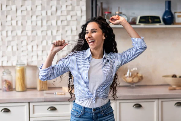 Mladá latinská žena se baví při vaření doma v kuchyni, drží whisk jako mikrofon, zpívá písně — Stock fotografie