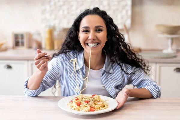 Happy excited latin woman eating tasty homemade pasta and smiling at camera, sitting at table in kitchen interior — Stock Photo, Image