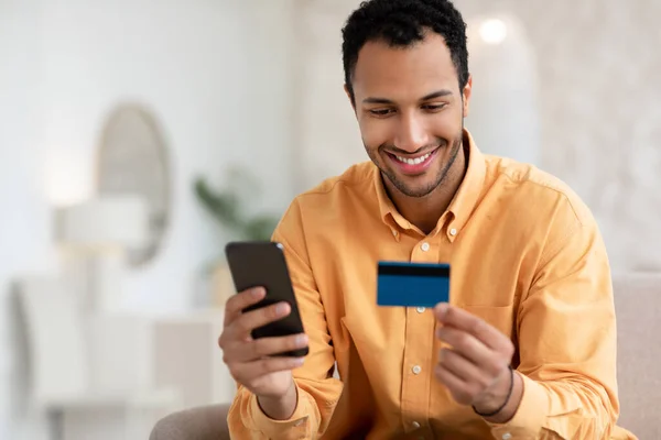 Sonriente chico árabe usando el teléfono celular y la tarjeta de crédito en casa —  Fotos de Stock