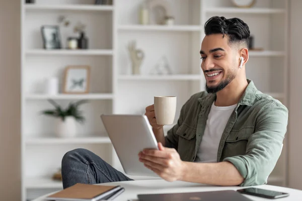 Sorrindo atraente jovem muçulmano macho com barba em fones de ouvido sem fio beber café, fazer chamada on-line — Fotografia de Stock