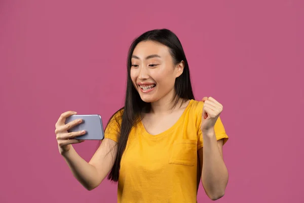 Yes, great news. Overjoyed korean lady celebrating win, holding cellphone and shaking clenched fist, violet background — Stock Photo, Image