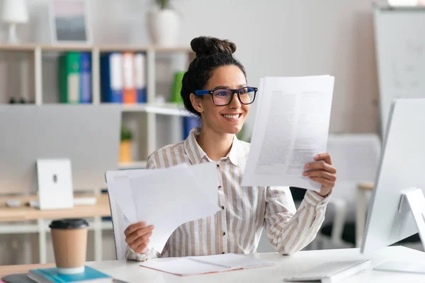 Feliz mujer de negocios latina mirando a través de documentos en su escritorio en la oficina, leyendo informes en su lugar de trabajo — Foto de Stock