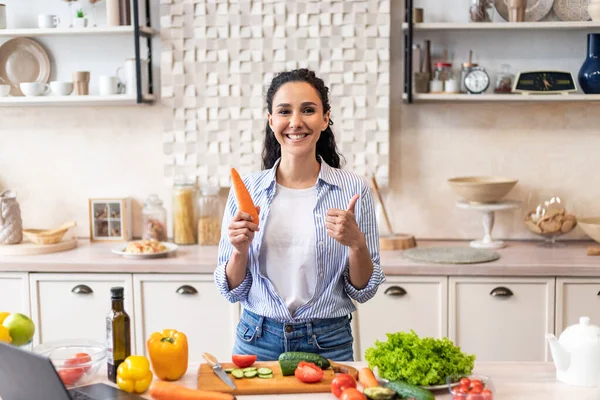 Счастливая латиноамериканка держит морковь и жестикулирует большим пальцем вверх, утверждая рецепт во время приготовления пищи на кухне — стоковое фото