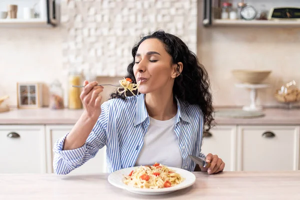 Young latin woman eating delicious pasta, enjoying tasty homemade lunch with closed eyes, sitting in kitchen — Stock Photo, Image