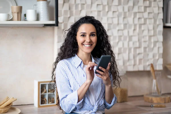 Feliz joven latina usando el teléfono inteligente mientras desayuna en el interior de la cocina en casa, sonriendo a la cámara — Foto de Stock