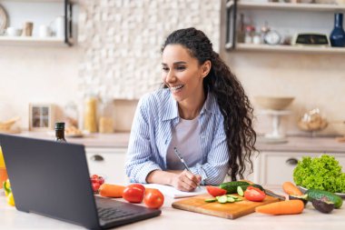 Yeni tarif yazıyorum. Mutlu Latin kadın laptopta webinar pişirirken defterine not alıyor.