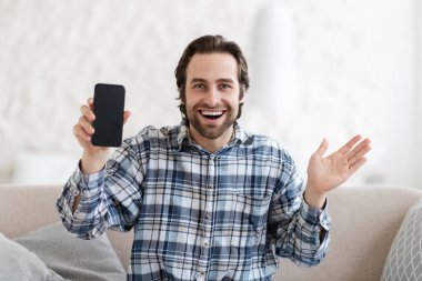 Tatmin olmuş, heyecanlı, gömlekli genç Avrupalı jest yapan, boş ekranlı telefonu kanepeye oturan,