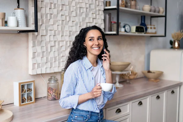 Glad latinamerikansk kvinna dricker kaffe och har telefonsamtal, står i köket interiör och ler — Stockfoto