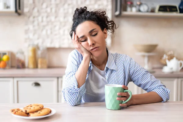 Schläfrige junge Frau sitzt mit geschlossenen Augen am Esstisch in der Küche und hält Tasse beim Frühstück — Stockfoto