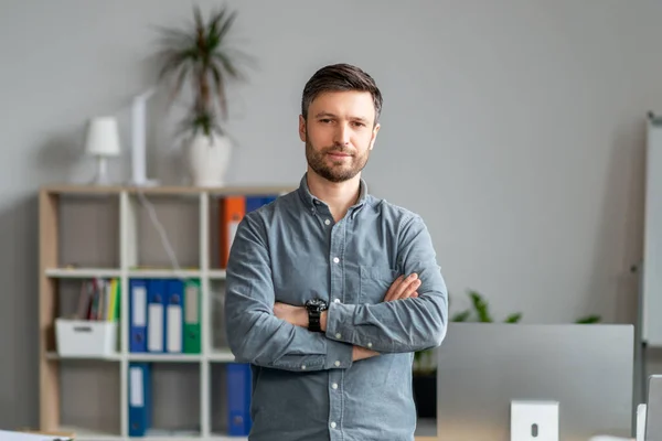 Porträtt av mogen affärsman står nära arbetsplatsen i ämbetet, poserar med korsade händer och tittar på kameran — Stockfoto