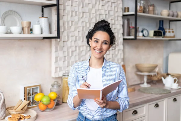 Mulher latina feliz segurando livro de culinária, escrevendo receita para o delicioso jantar, de pé na cozinha e sorrindo para a câmera — Fotografia de Stock