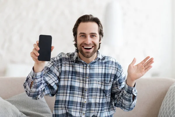 Satisfecho sorprendido joven europeo excitado en el gesto de la camisa, mostrar el teléfono con pantalla en blanco sentado en el sofá — Foto de Stock