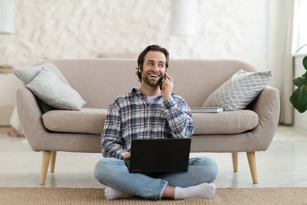 Feliz millennial europeo chico con rastrojo en las llamadas de camisa por teléfono, funciona en el ordenador portátil, se sienta en el suelo en la sala de estar — Foto de Stock