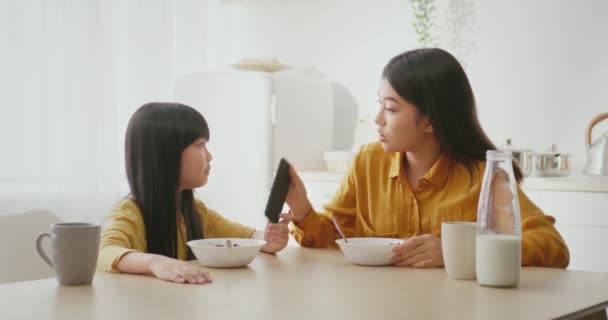 Kids and gadgets. Cute little asian girl eating breakfast and networking on smartphone, mother scolding her at kitchen — Stock Video
