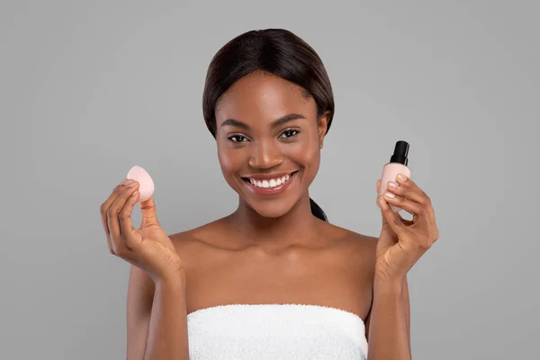 Mulher negra alegre em toalha fazendo maquiagem básica com cosméticos, segure esponja e corretor, isolado em fundo cinza — Fotografia de Stock