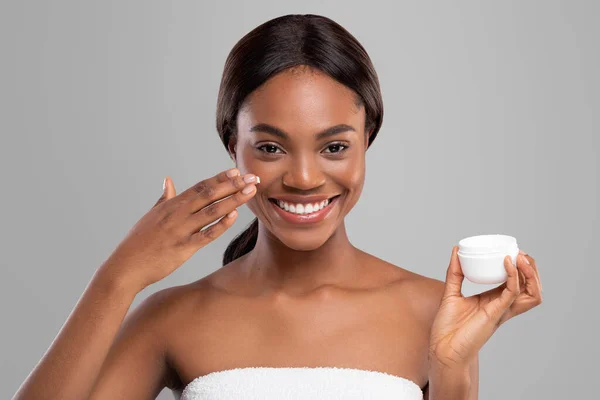 Feliz joven mujer negra con la piel perfecta aplicando crema hidratante anti-envejecimiento en su cara y sostener el frasco —  Fotos de Stock