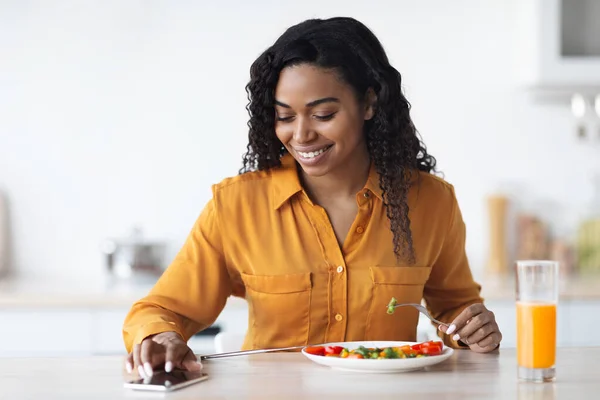 Glada afrikanska amerikanska kvinna som äter lunch, med hjälp av smartphone — Stockfoto