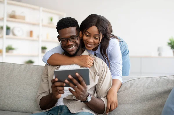 Vrolijk Afro-Amerikaans echtpaar zittend op de bank, met behulp van tablet — Stockfoto
