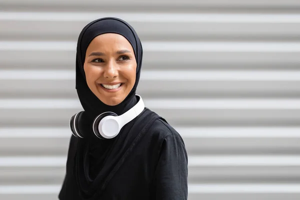 Glada unga arabiska dam idrottsman i hijab och trådlösa hörlurar titta på tomt utrymme på väggen bakgrund — Stockfoto