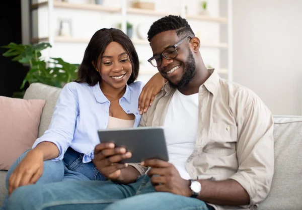 Cheerful African American couple sitting on sofa, using tablet — ストック写真