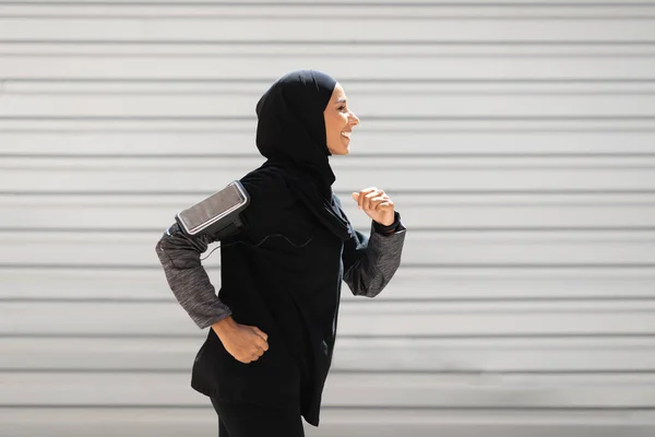 Happy young islamic athlete in hijab, sportswear with smartphone runs on wall background, profile — Stok Foto
