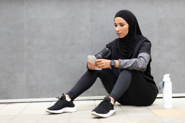 Kendine güvenen, tesettürlü genç Arap bayan sporcu, telefon ve su şişesiyle spor giyim, gri duvar arkasında oturuyor.