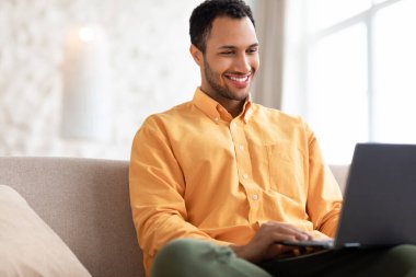 Arap adam evdeki koltukta dizüstü bilgisayar kullanıyor.