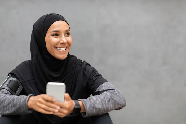 Senyum muda muslim atlet wanita di jilbab pakaian olahraga dengan smartphone melihat ruang kosong — Stok Foto