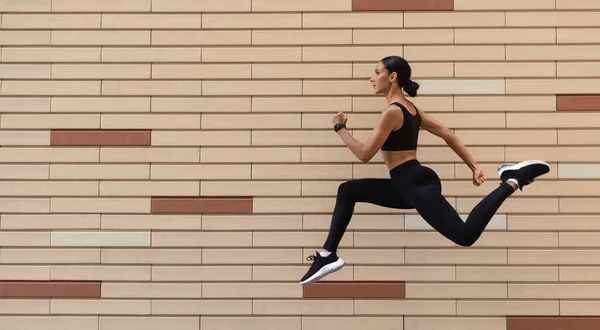 Joven atleta árabe seria en carreras de ropa deportiva, congelado en el aire sobre fondo de pared de ladrillo, perfil, panorama —  Fotos de Stock