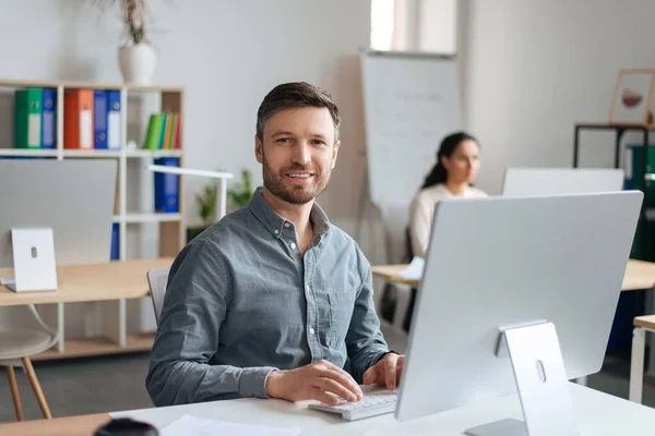 Schöner, fröhlicher Mann sitzt am Schreibtisch und benutzt einen modernen Computer im Großraumbüro — Stockfoto