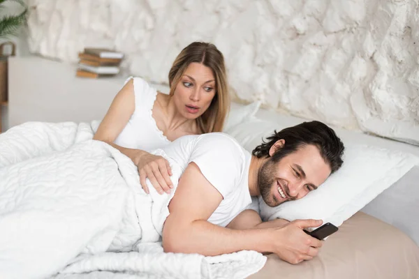 Zszokowana młoda kobieta widząc męża wysyłającego wiadomość do kochanka na smartfonie leżącego w łóżku w domu — Zdjęcie stockowe
