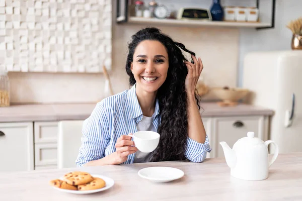 Portret van vrolijke latijn langharige vrouw aan keukentafel, genietend van 's morgens warme drank thuis, kopieerruimte — Stockfoto
